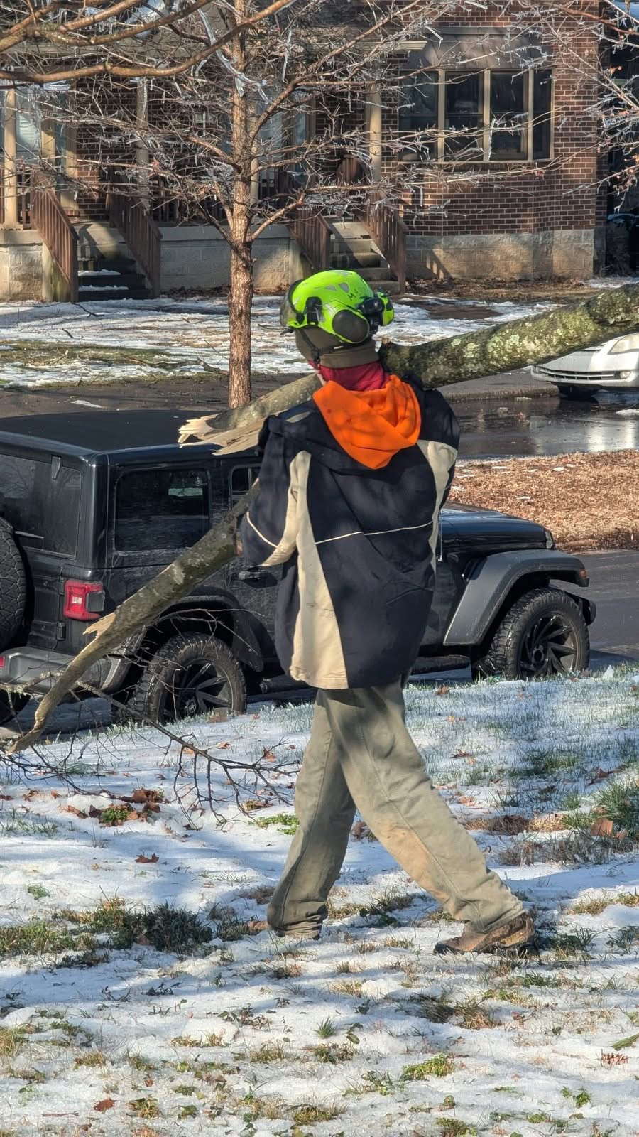 Crew member in a helmet shouldering a long splintered limb across a snow-patched yard, ice-covered branches in the background