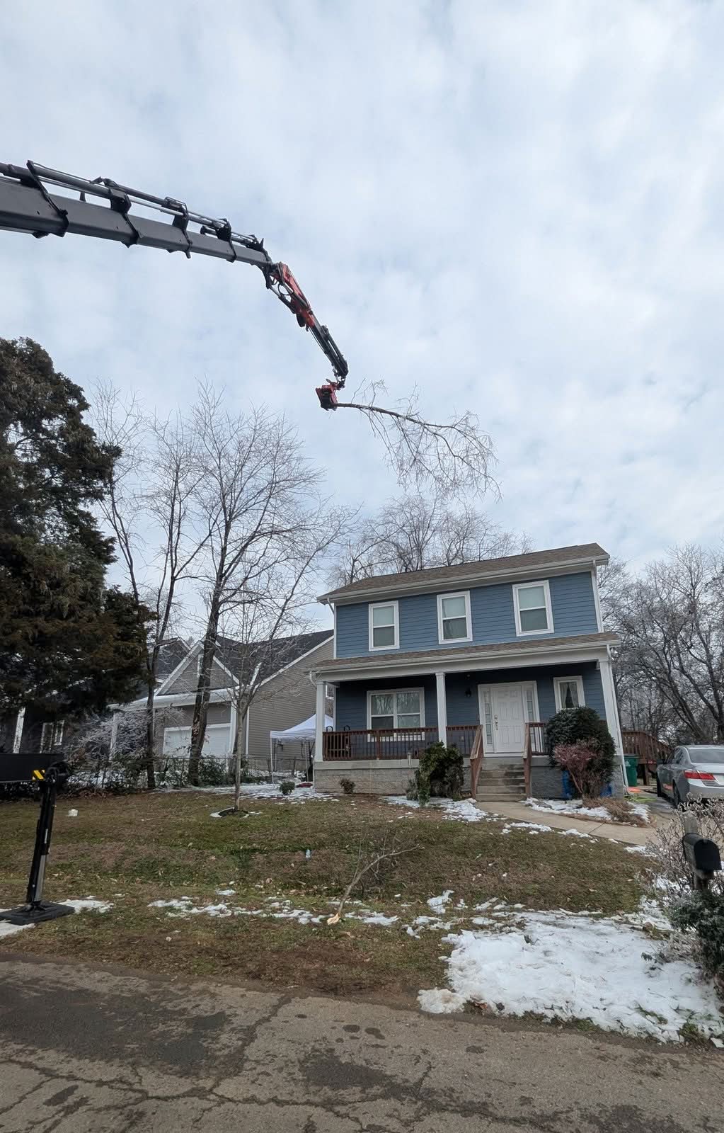 Grapple-saw crane carrying a cut branch high above a two-story house, patchy snow on the lawn below