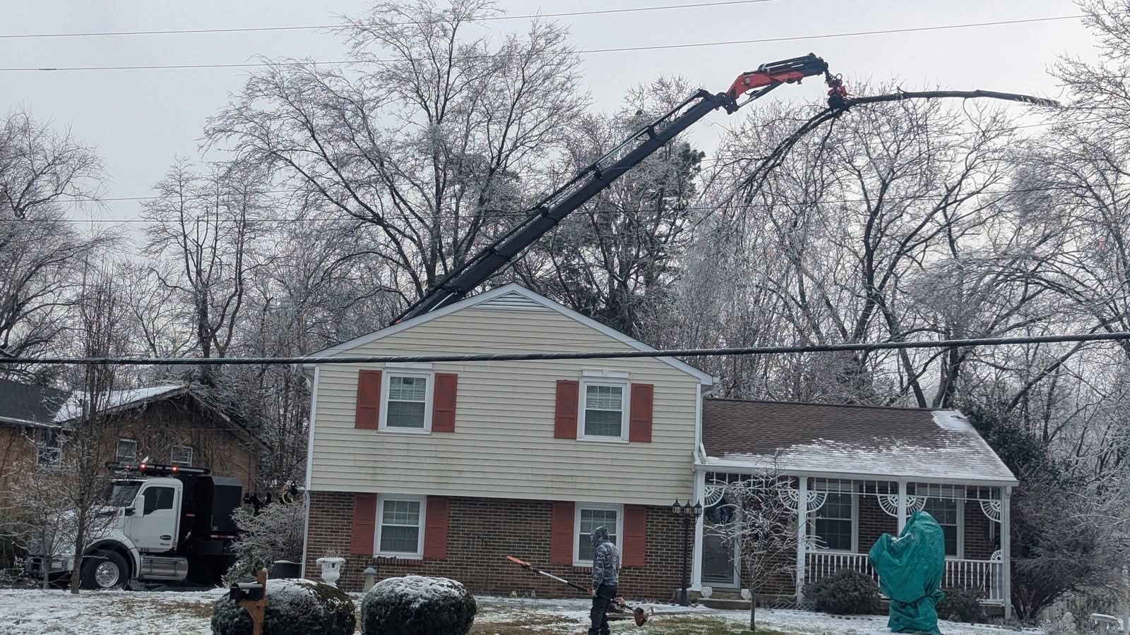 Red Palfinger knuckleboom crane parked in front of a house, boom arched over the roof to grip a limb in ice-covered trees