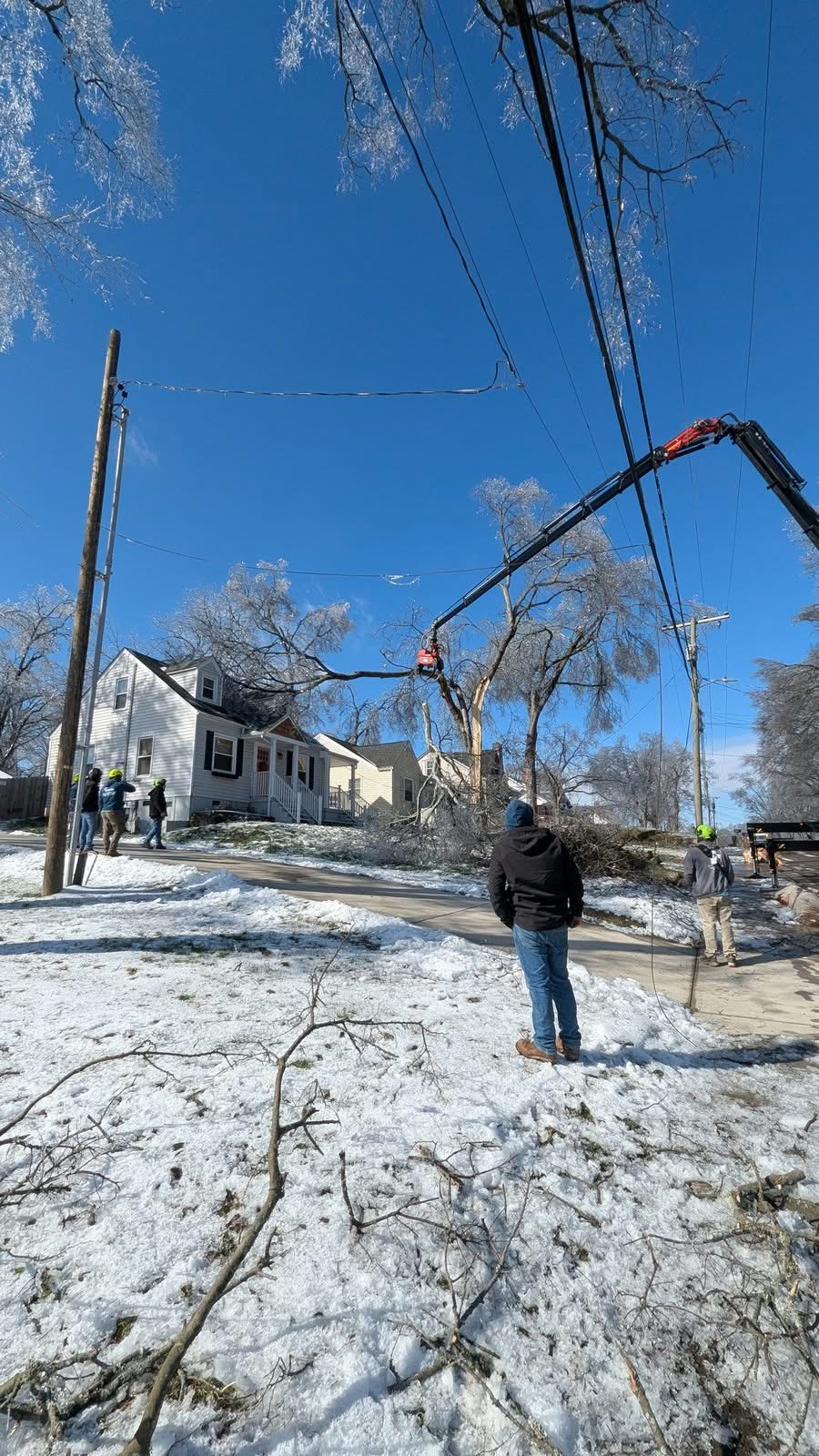 Knuckleboom crane boom extended over a snowy street toward a split, ice-coated tree, crew standing back near the power lines