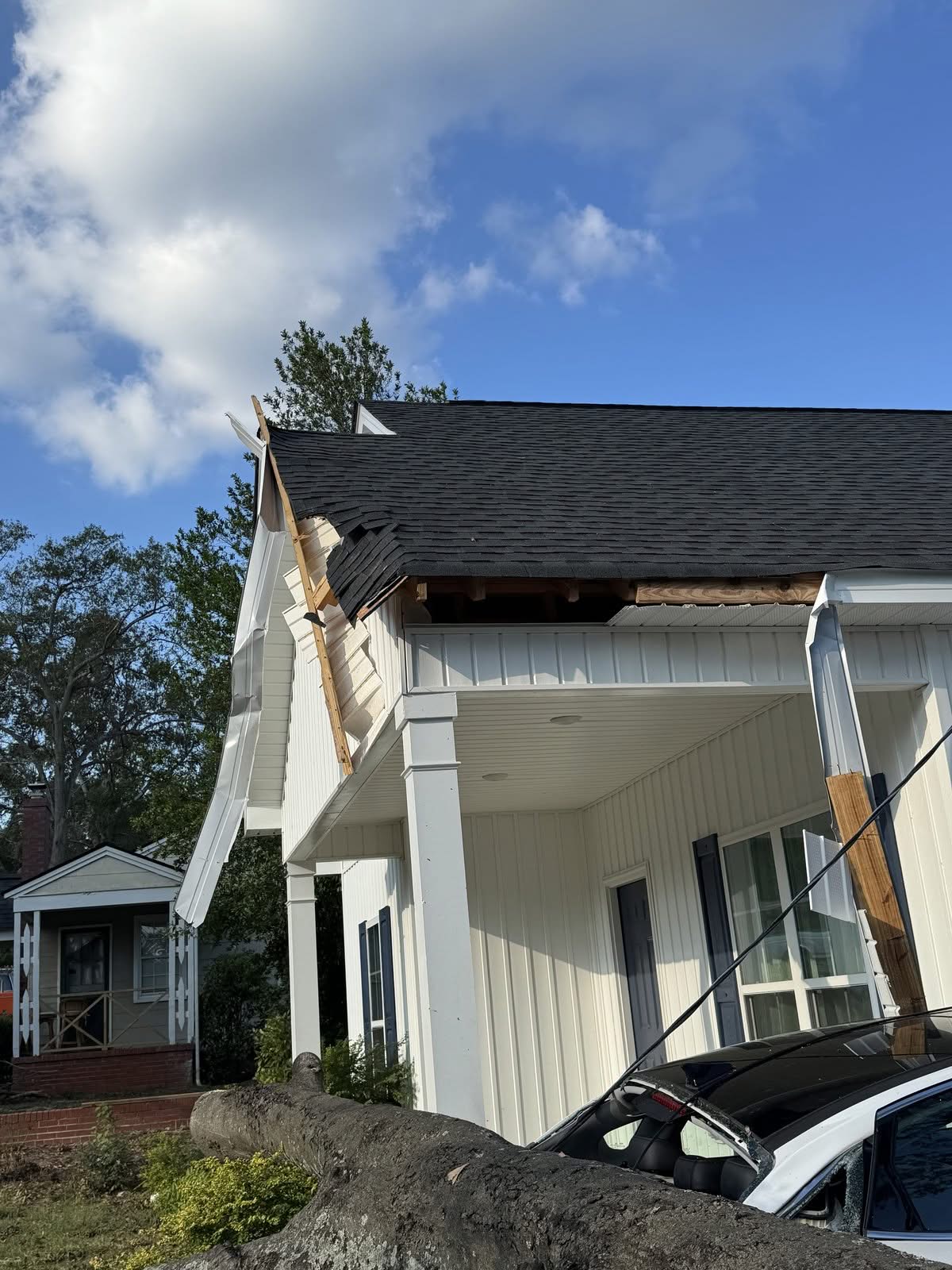 Tree trunk resting on a crushed car beside a torn porch roof after a storm